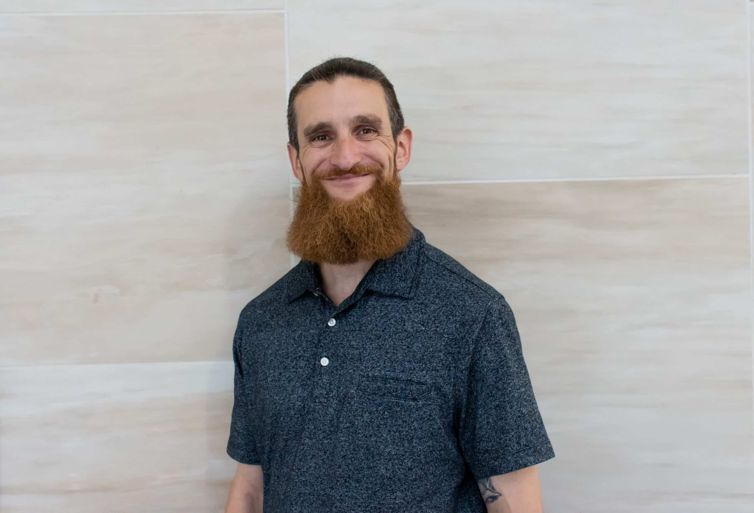 man with dark hair and a full long beard, wearing a blackand gray speckled polo shirt.