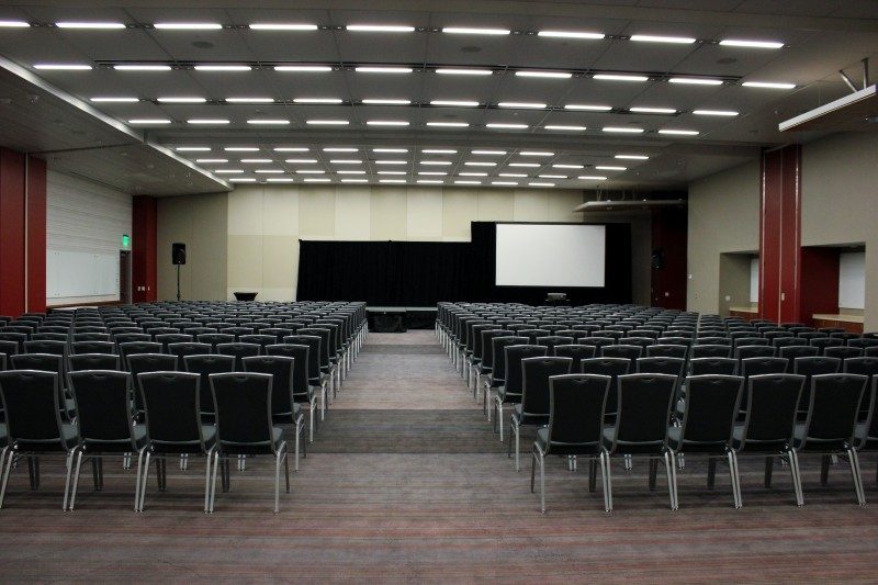 hundreds of chairs facing a stage with black drape behind it as well as a tall projection screen lined with black drape.