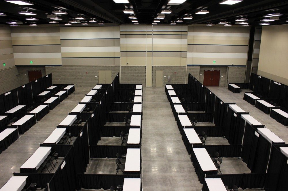 trade show booths consisting of black drape, one table, and two chairs set in a large room with high ceilings. The ceiling is black, the walls have blue, white, and tan stripes. The floor is polished gray concrete