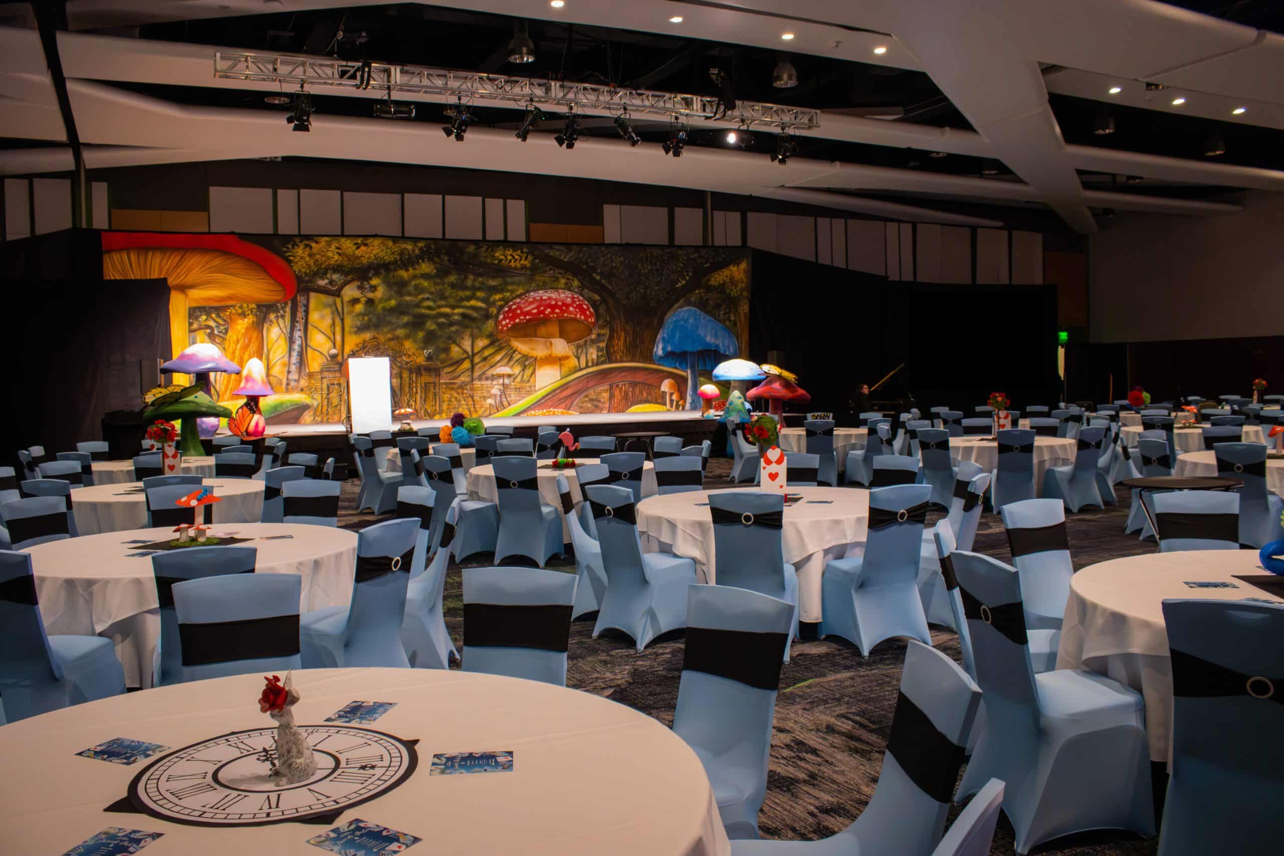 Room decorated with an Alice in Wonderland theme--mushroom backdrop across stage, playing cards on tables, tables set with white linens. chairs covered with white chaircovers and black sashes