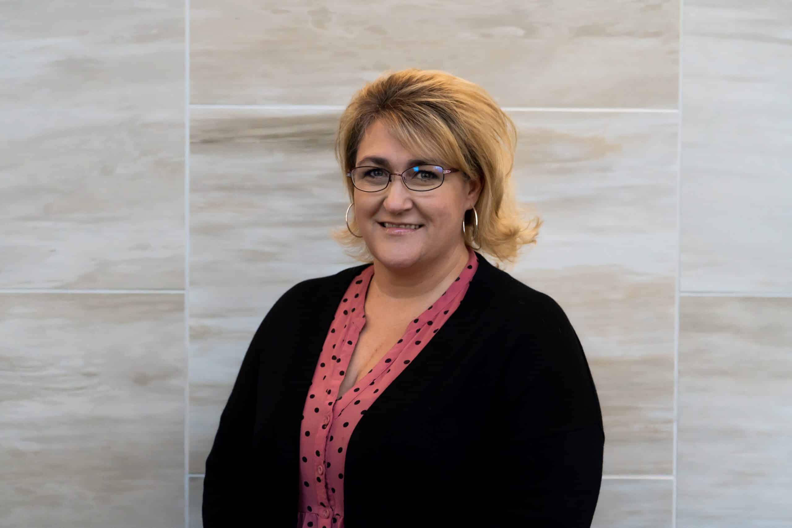 woman with short, blonde hair wearing earrings, glasses, a pink shirt with black dots and a black sweater